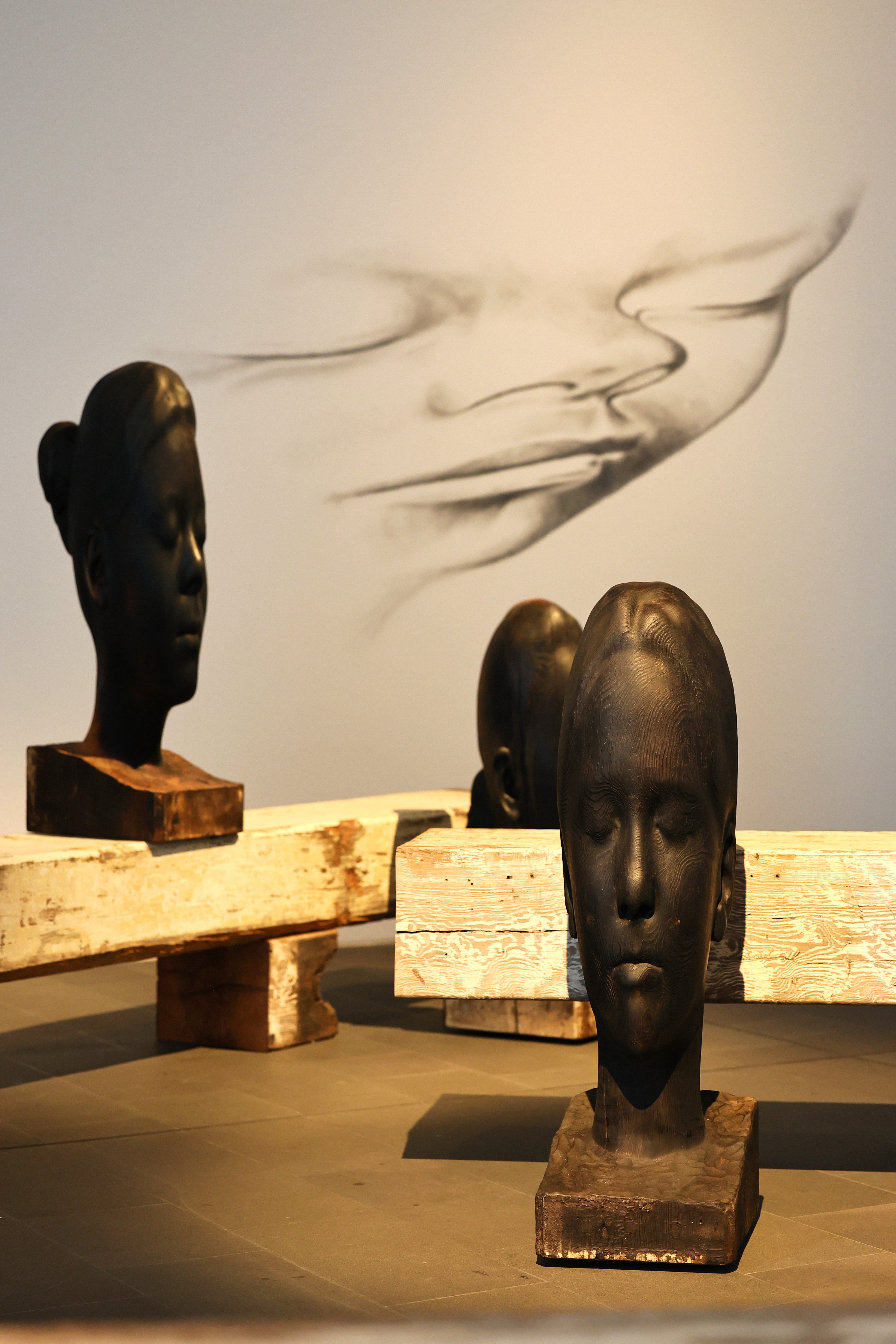 Köpfe zwischen Material und Transparenz Mehrere kleinere Kopfskulpturen stehen in einem Ausstellungsraum neben einer transparent wirkenden, geschwungenen Form von Jaume Plensa.
