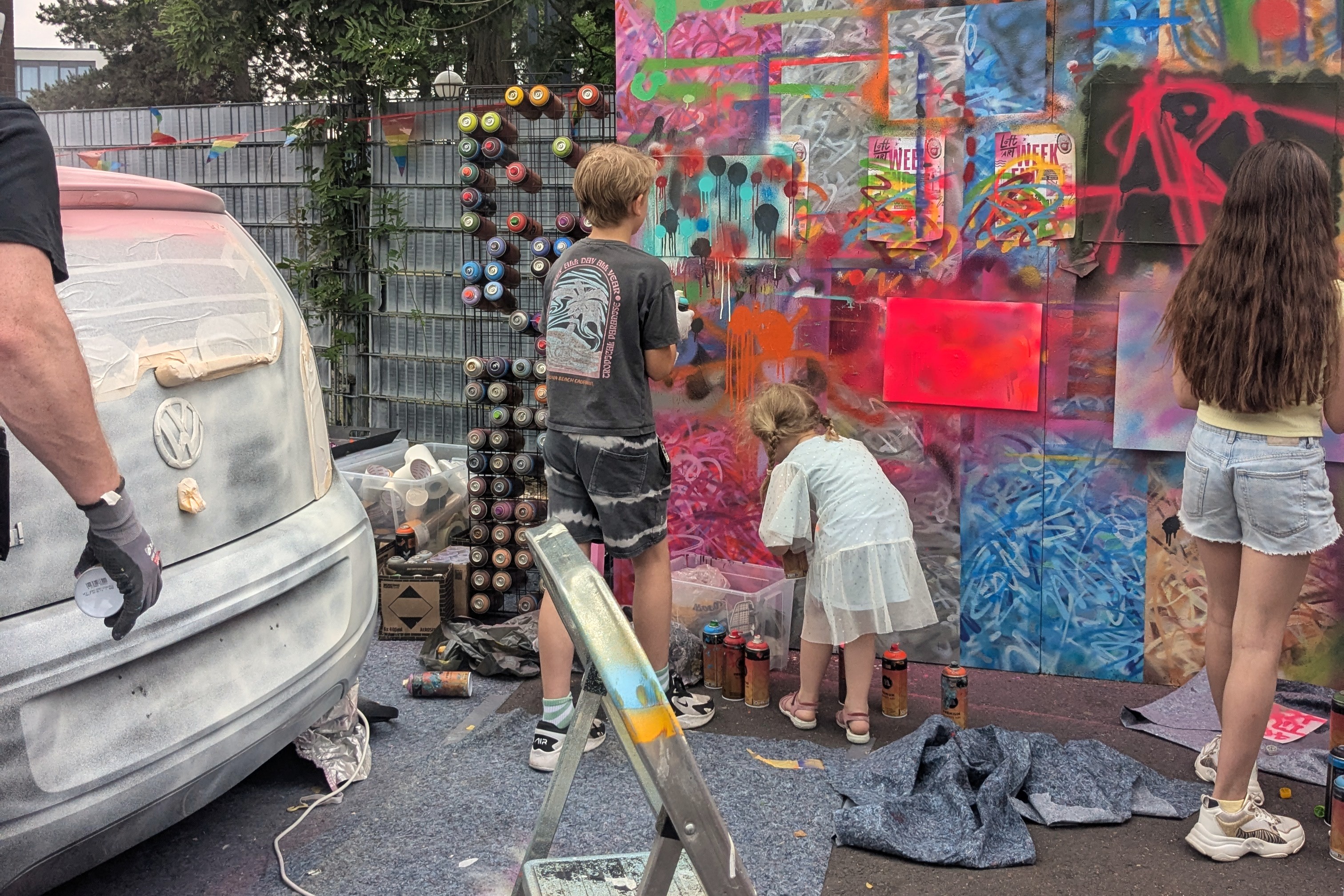 Zwei Kinder sprühen mit Spraydosen an einer bunten Wand, daneben ein mit Farbe besprühtes Auto und eine Halterung voller Spraydosen.