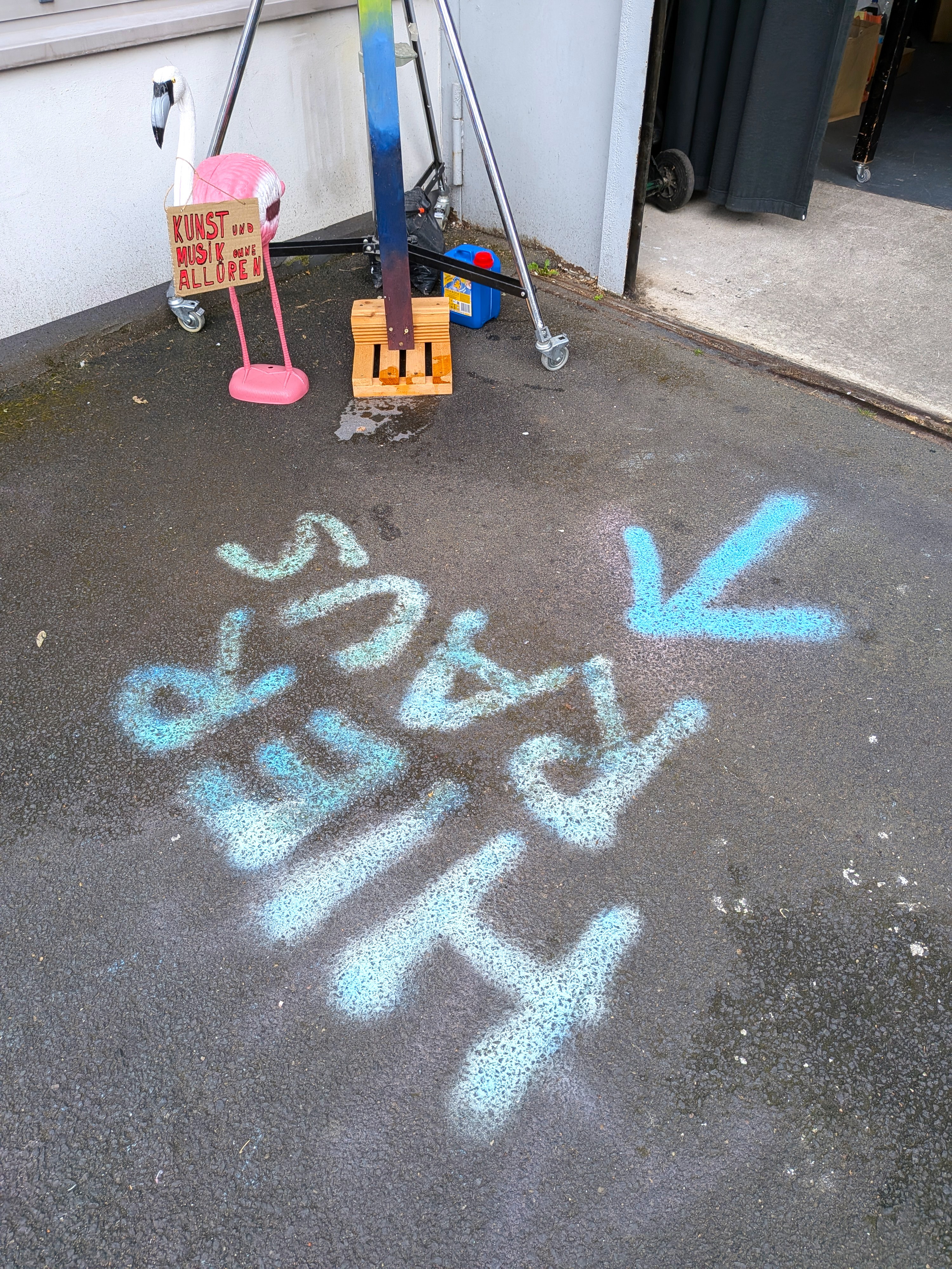Eine pinke Flamingo-Figur trägt ein Pappschild mit der Aufschrift Kunst und Musik ohne Allüren, auf dem Asphalt davor ein blaues Sprühbild.