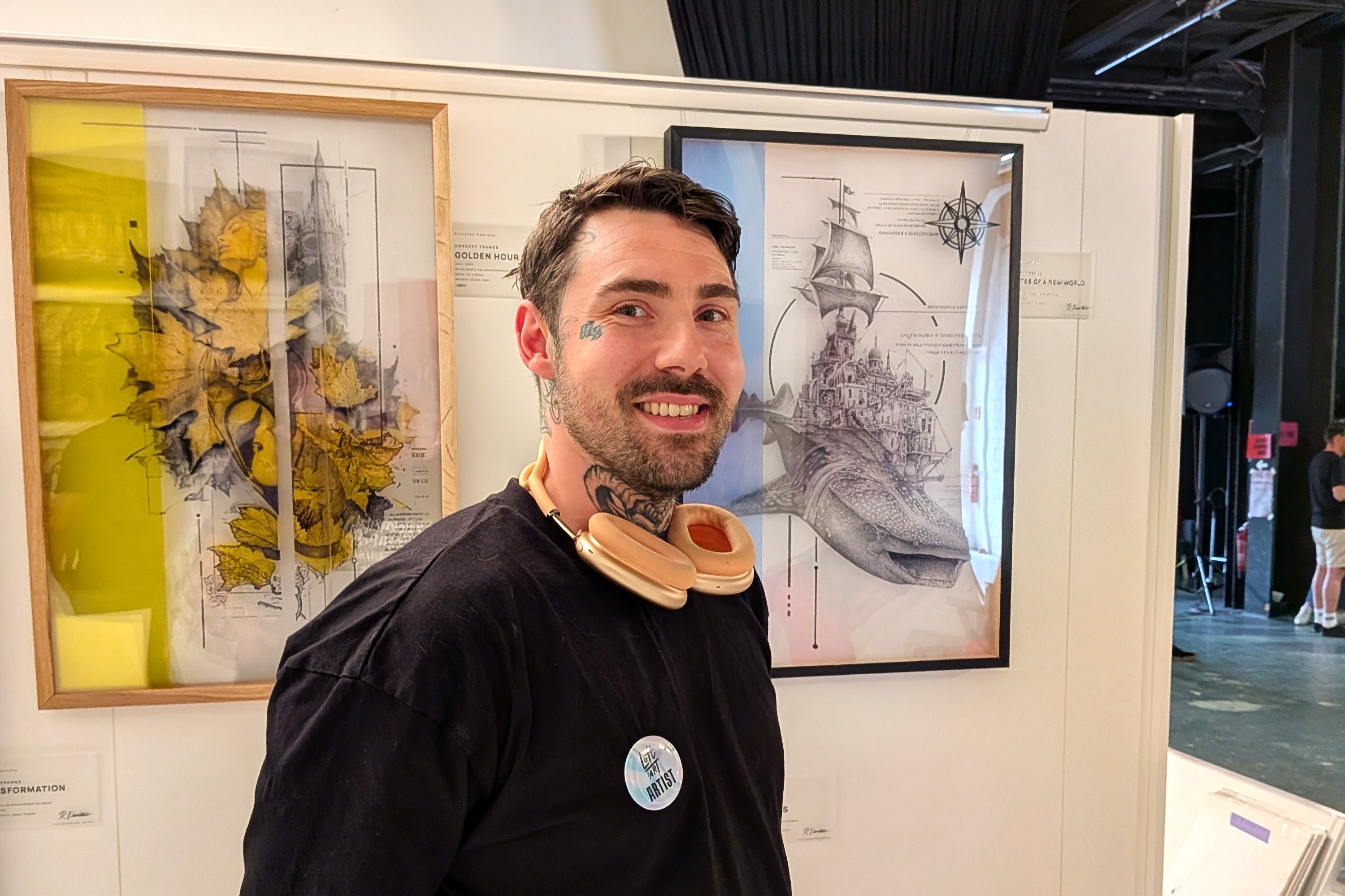 Ein tätowierter Mann mit Kopfhörern um den Hals steht vor zwei gerahmten Mixed-Media-Arbeiten, rechts eine Zeichnung von Schiff und Wal, daneben eine Künstlerkarte mit dem Namen Riccardo Damiano.