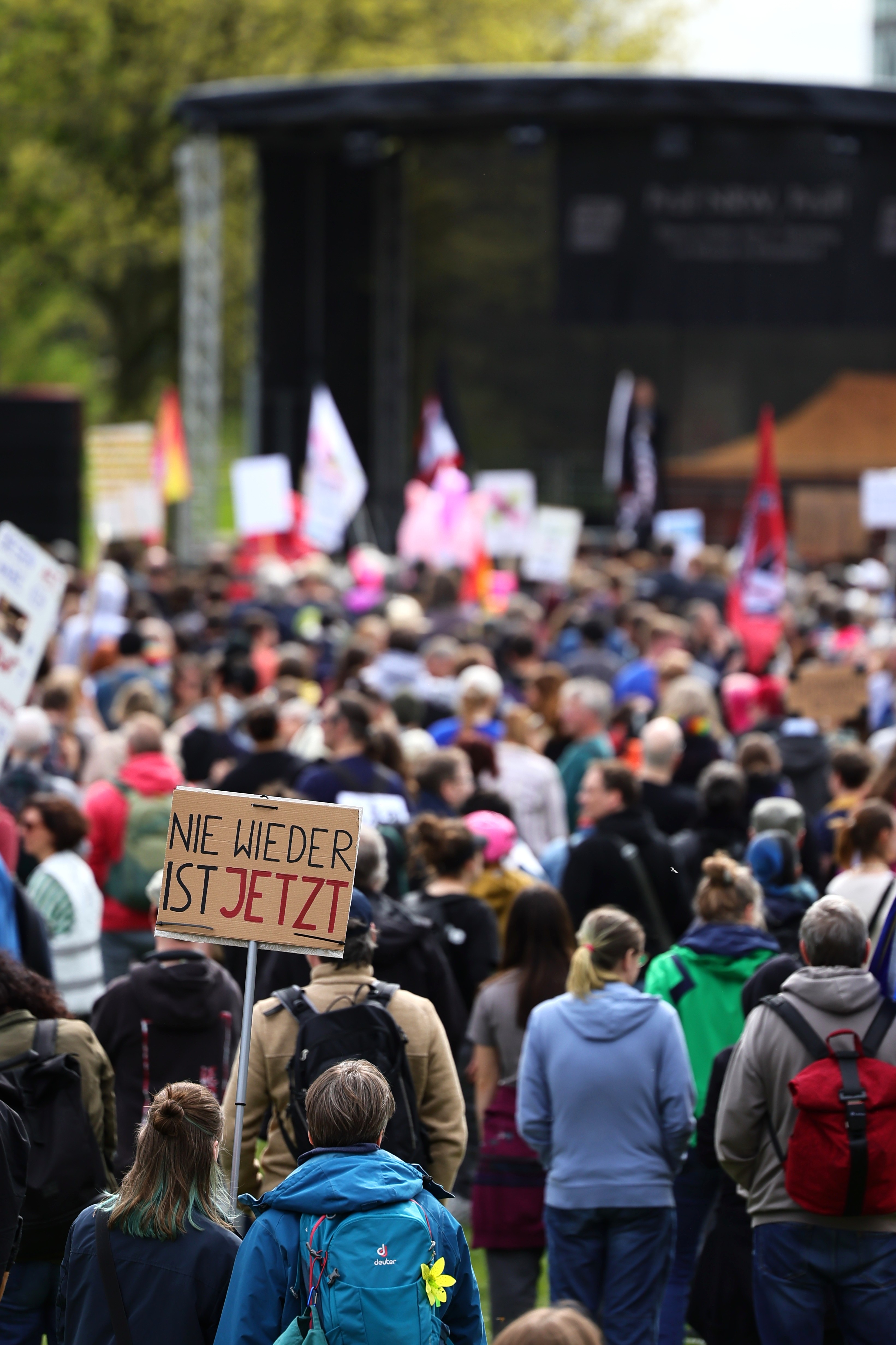 Demonstrant hält Schild „Nie wieder ist jetzt" inmitten der Menge auf der Landtagswiese bei der PRÜF-Demo Düsseldorf April 2026
