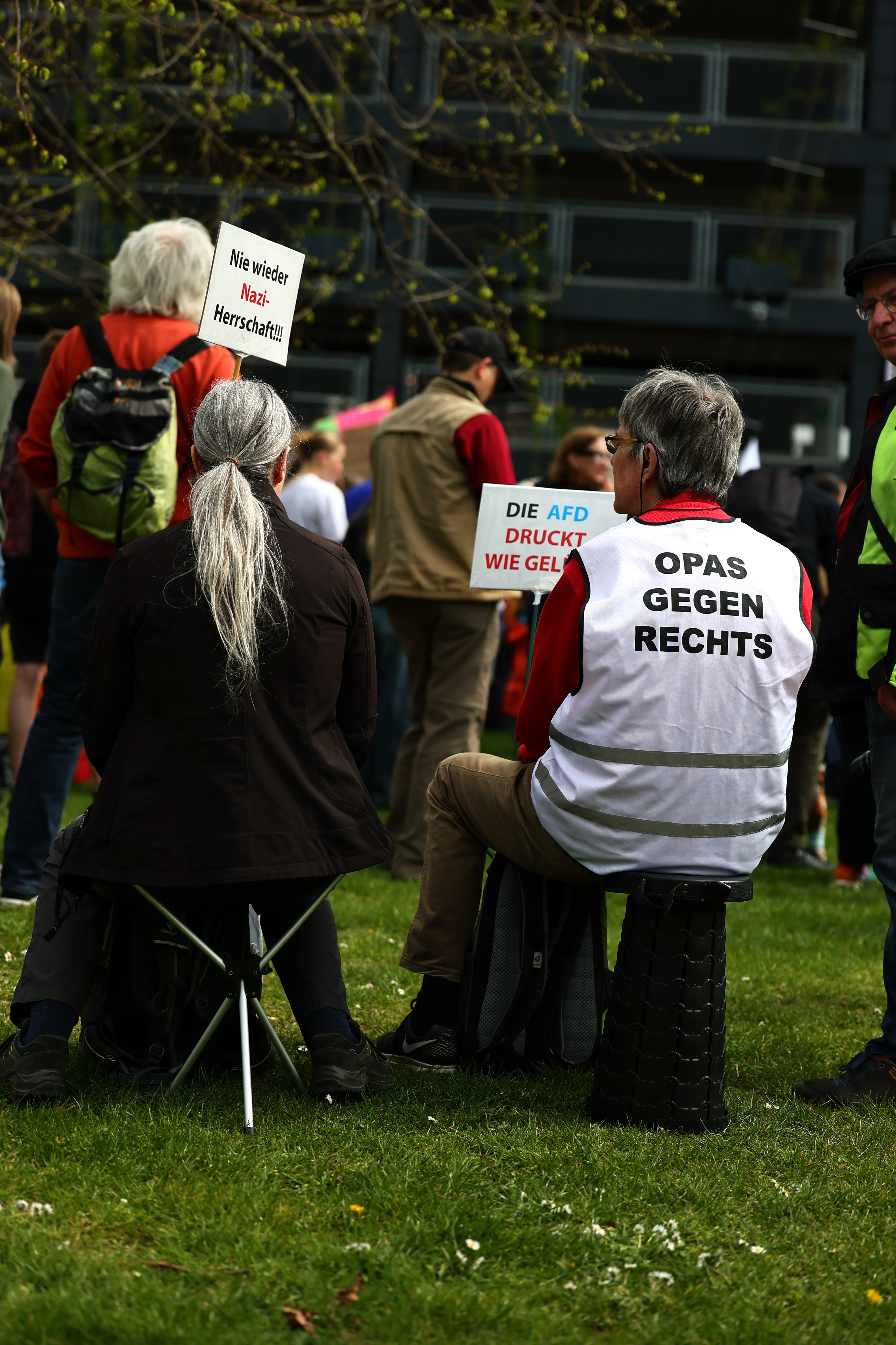Zwei ältere Menschen sitzen auf der Landtagswiese, einer trägt eine Weste mit der Aufschrift „Opas gegen Rechts" bei der PRÜF-Demo Düsseldorf