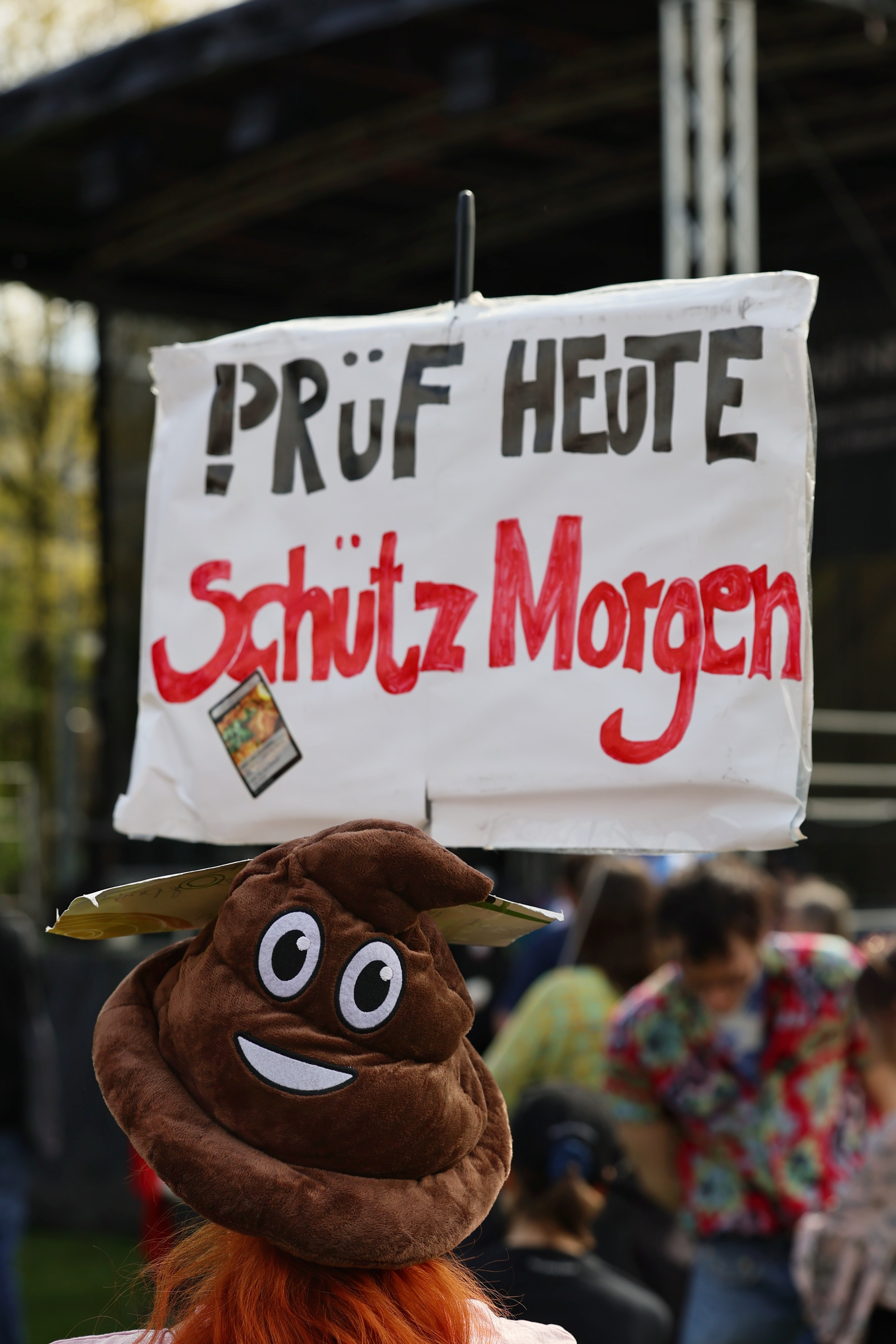 Person mit Kot-Emoji-Plüschmütze hält Schild mit Aufschrift „PRÜF heute – Schütz Morgen" bei der PRÜF-Demo auf der Landtagswiese Düsseldorf