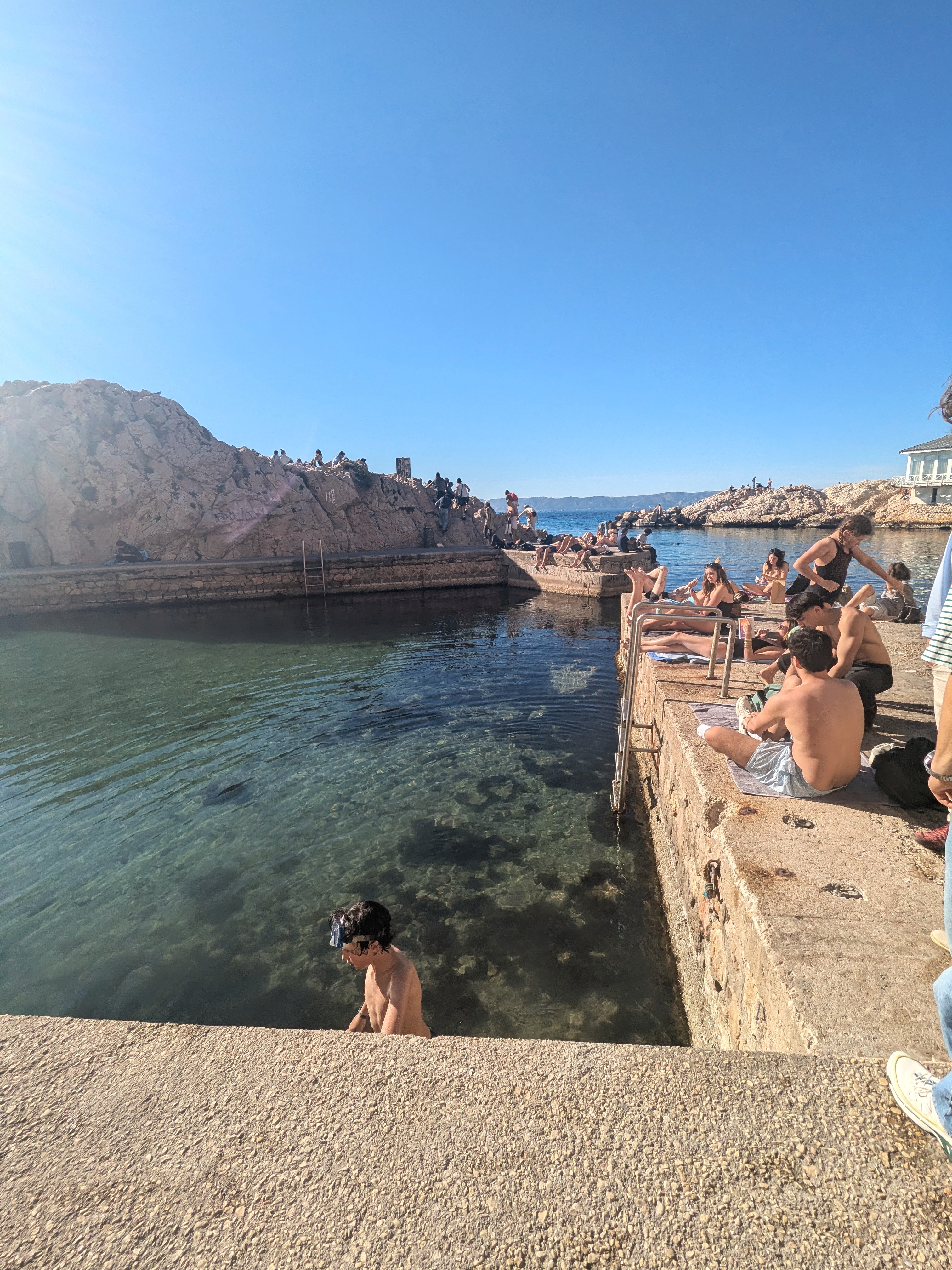 Junge mit Tauchermaske steigt aus dem Wasser an der natürlichen Badestelle am Vallon des Auffes in Marseille, Jugendliche sitzen auf den Felsen dahinter