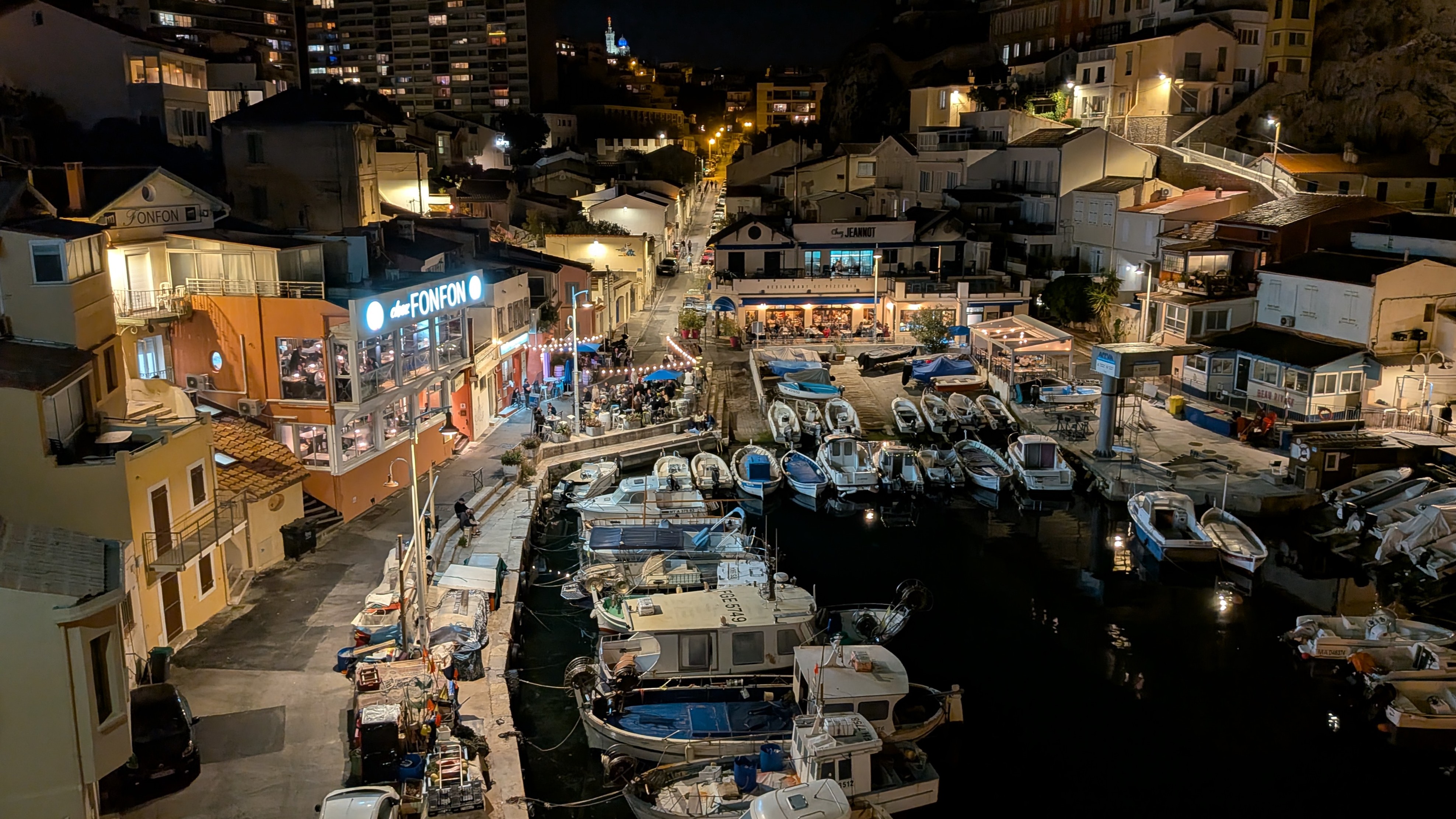Vallon des Auffes in Marseille bei Nacht: kleiner Fischerhafen mit beleuchtetem Restaurant Chez Fonfon, Booten und Notre-Dame de la Garde im Hintergrund