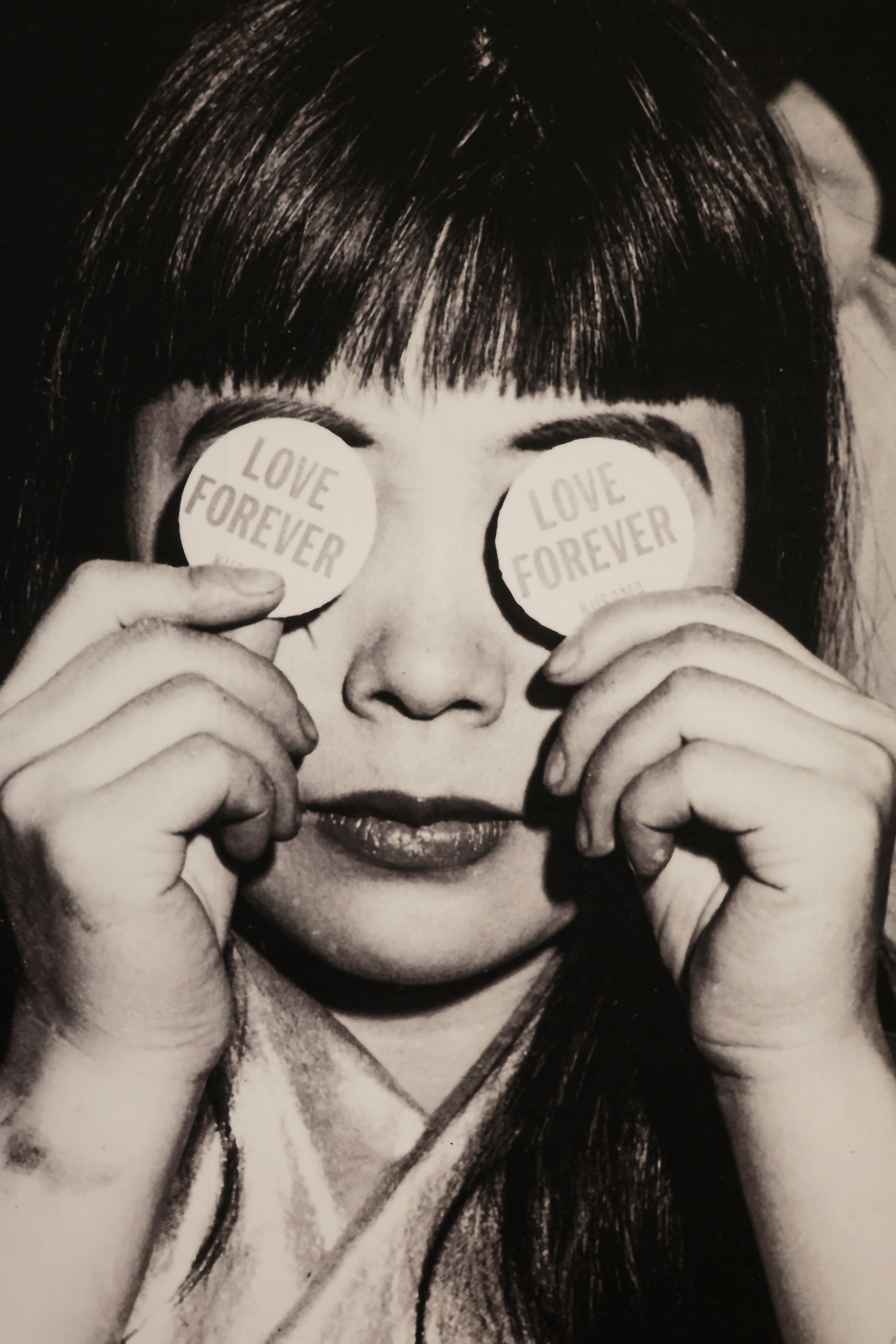 Schwarz-Weiß-Archivporträt von Yayoi Kusama: Die Künstlerin hält zwei runde Buttons mit der Aufschrift „Love Forever" vor ihre Augen